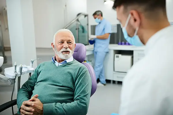 An older male patient sitting in a dental chair and consulting with a dentist about the dangers of dry mouth.